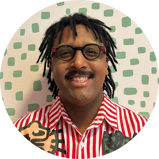 Mateus: pessoa negra com blusa listrada colorida, dreads escuros e óculos